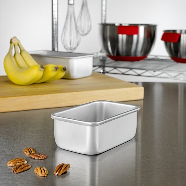 A silver Fat Daddio's bread loaf pan on a counter with bananas and nuts.