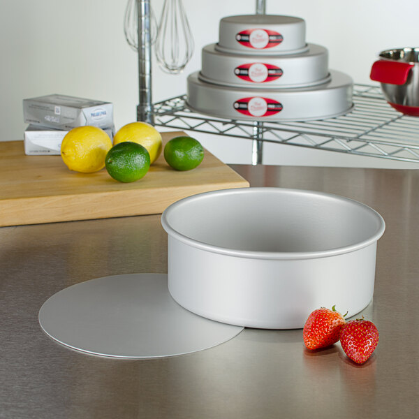 A Fat Daddio's round metal cake pan with a red lid on a counter with fruit.