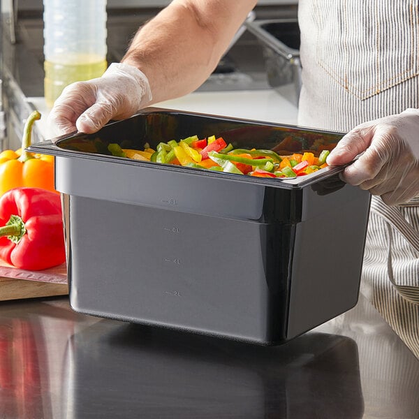 A person in gloves holding a black Vigor 1/2 size polycarbonate food pan filled with vegetables.