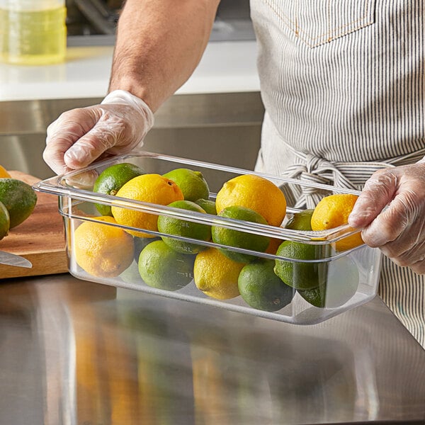 A person in a white shirt and gloves holding a Vigor clear polycarbonate food pan of lemons and limes.
