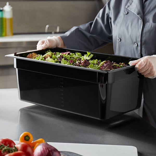 A person in a chef's uniform holding a black Vigor food pan filled with food on a counter in a salad bar.