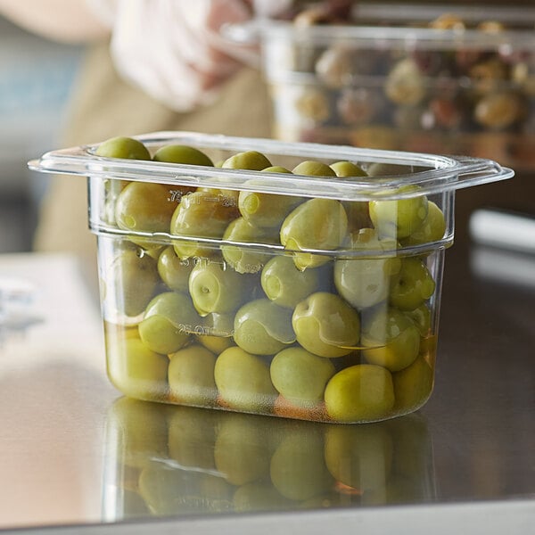A person using a Vigor 1/9 size clear polycarbonate food pan to hold green olives on a counter.