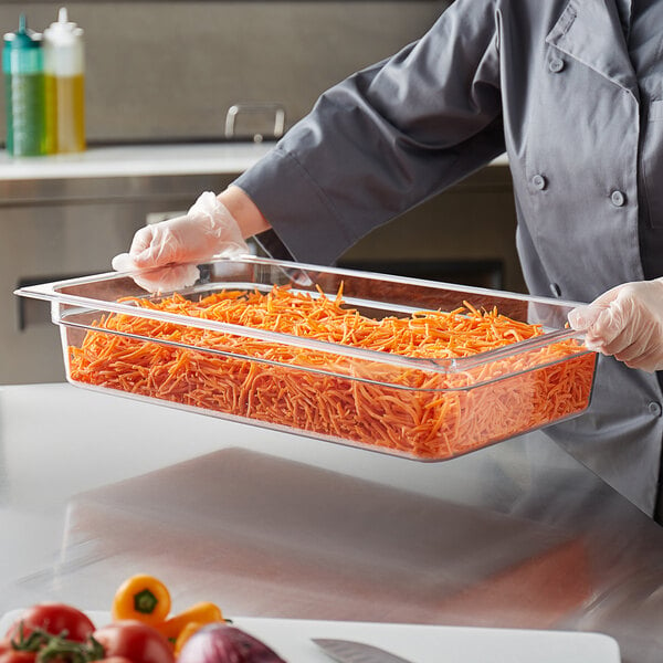 A person in a chef's uniform holding a clear polycarbonate food pan of shredded carrots.