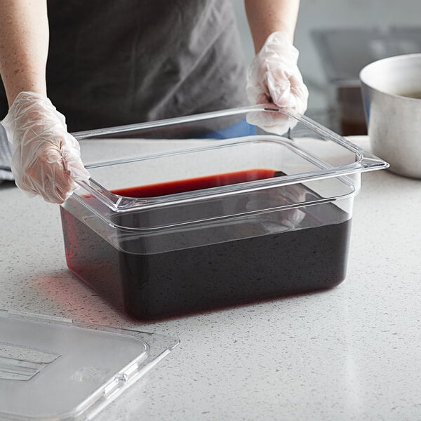 A person in gloves holding a Vigor 1/2 size clear polycarbonate food pan filled with red liquid.