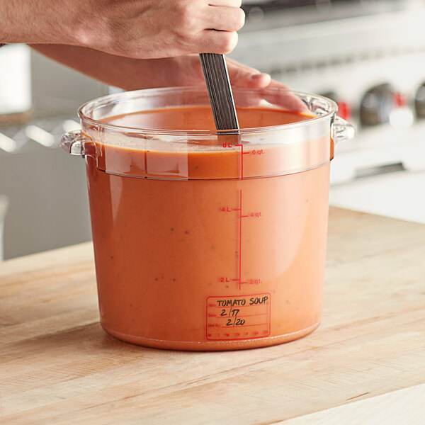 A person stirring tomato soup in a large clear plastic container.