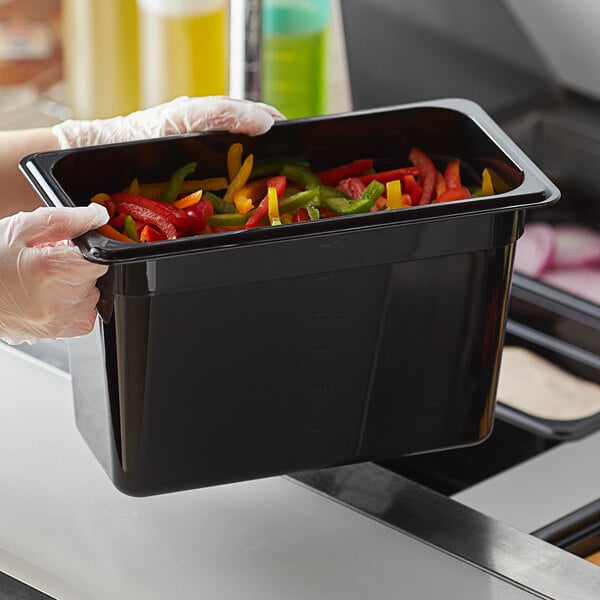 A person in gloves holding a black Vigor polycarbonate food pan filled with peppers.