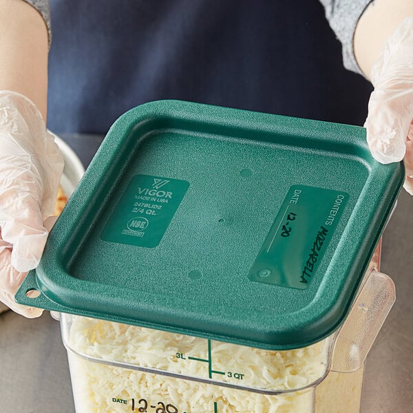 A person in gloves holding a Vigor square green polypropylene food storage container filled with shredded cheese.