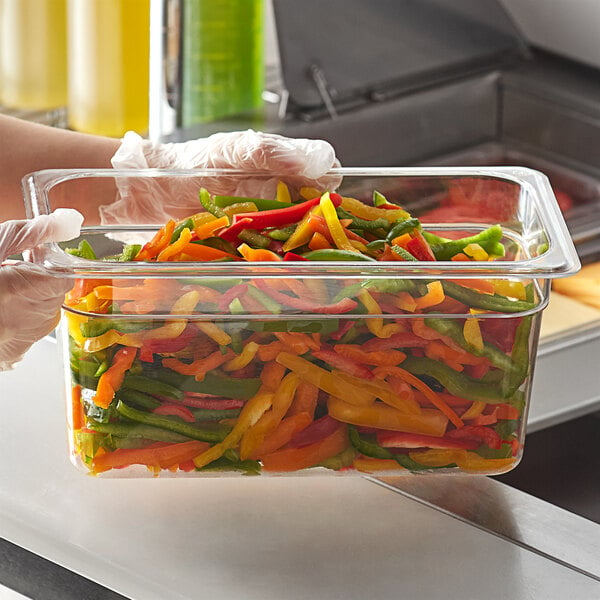 A person in gloves holding a clear polycarbonate food pan of colorful peppers.