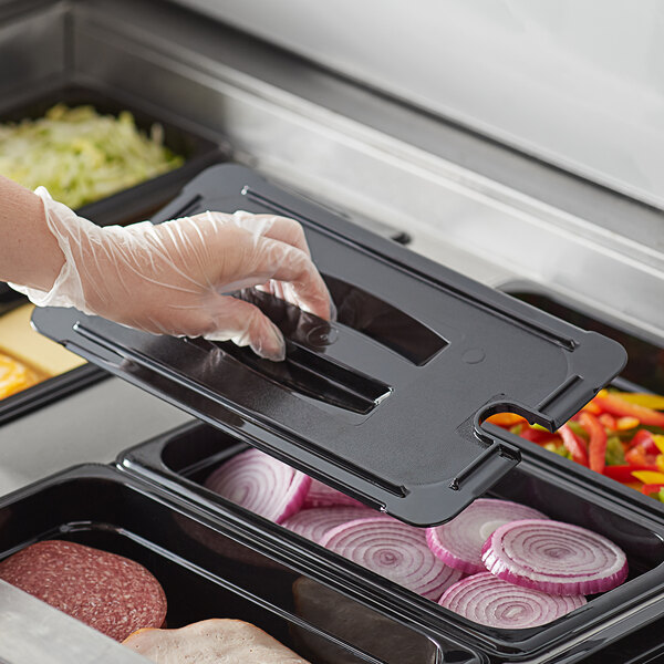 A person wearing plastic gloves puts food into a tray using a black Vigor polycarbonate food pan lid with a handle and notch.