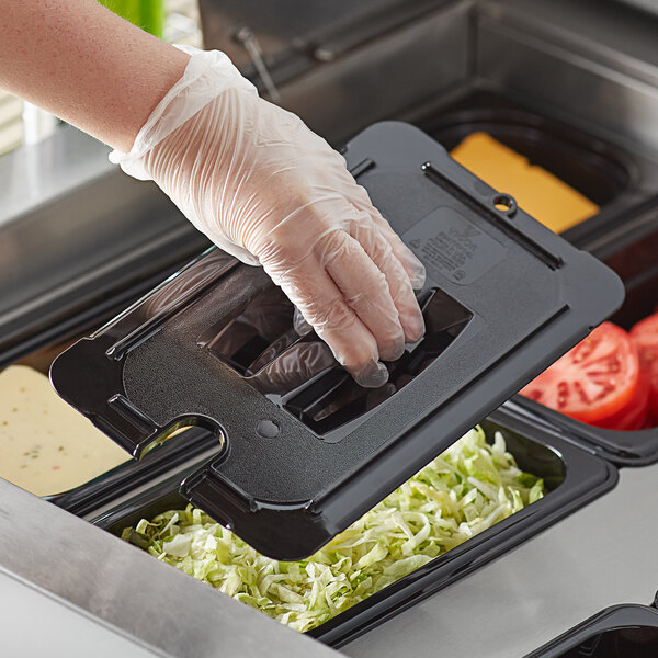 A hand in a glove holding a black Vigor polycarbonate food container lid with food in it.