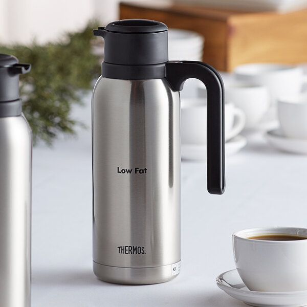 A silver and black Thermos stainless steel vacuum insulated carafe on a white table.