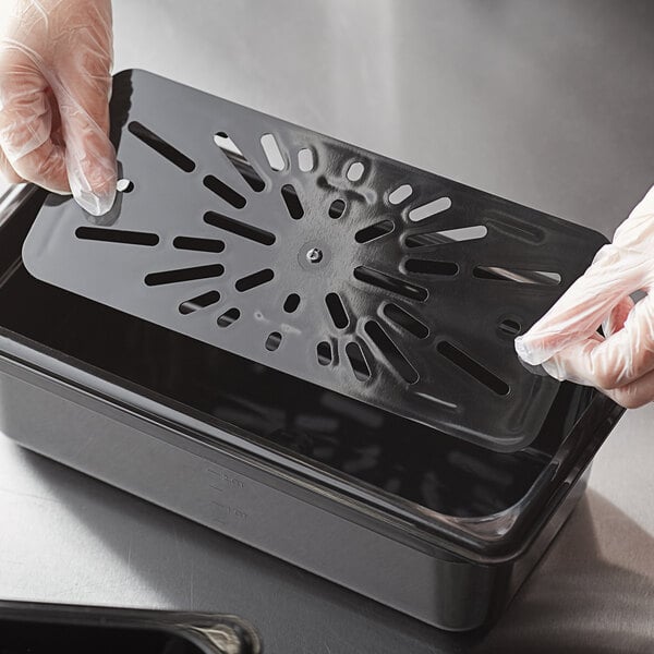 A gloved hand placing a black Vigor polycarbonate drain tray in a plastic container on a counter.