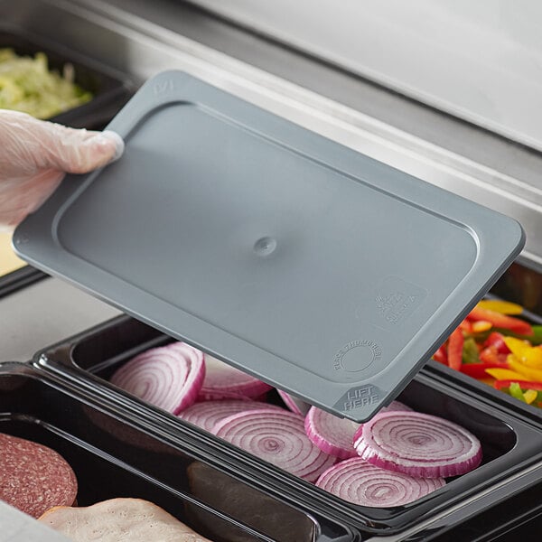 A person holding a Vigor 1/3 size gray plastic food pan cover over a tray of food.