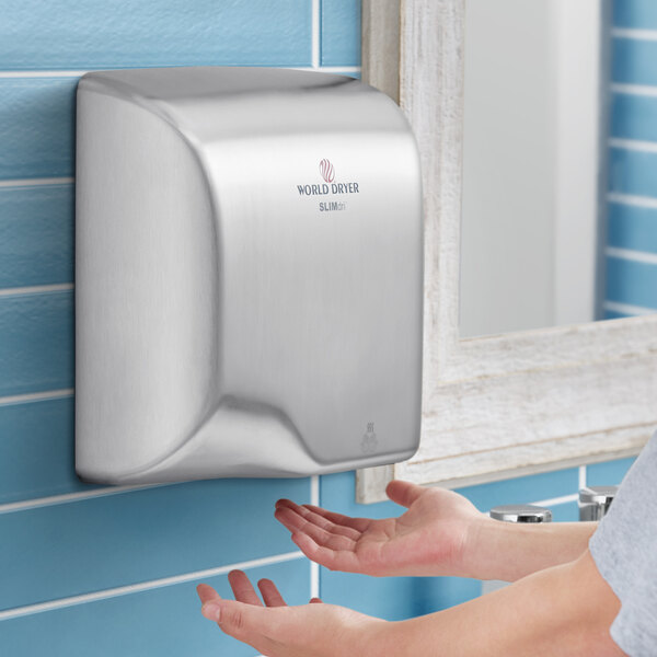 A person using a World Dryer brushed stainless steel surface-mounted hand dryer.