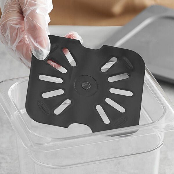 A plastic gloved hand holding a black Vigor polycarbonate drain tray in a plastic container.