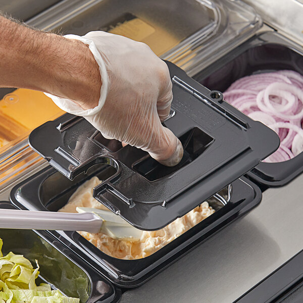 A hand in a plastic glove using a black Vigor polycarbonate food pan lid to cover a container of food.
