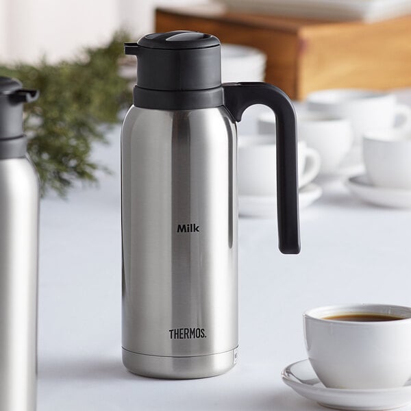 A silver and black stainless steel vacuum insulated carafe by Thermos on a white table.