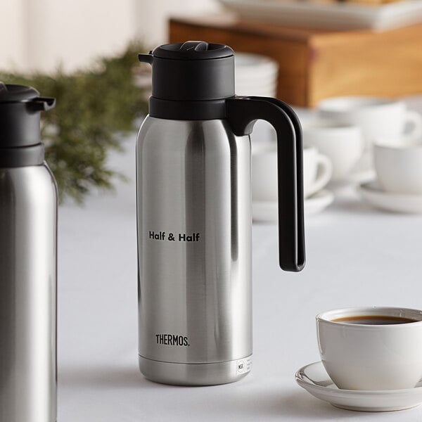 A silver and black Thermos carafe on a table with a cup of coffee.