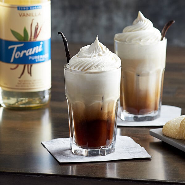 Two glasses of coffee with whipped cream and vanilla sticks with a bottle of Torani Puremade Zero Sugar Vanilla Syrup.