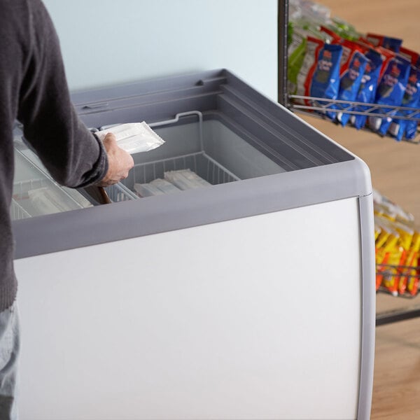 A person in a blue shirt opening an Avantco flat top display ice cream freezer and putting a bag of food inside.