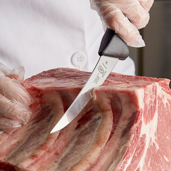 A gloved hand uses a Mercer Stiff Curved Boning Knife to cut a piece of meat.