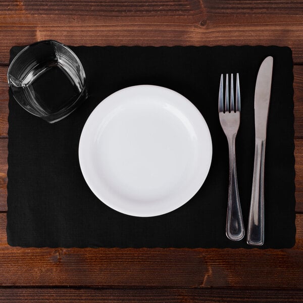 A white plate, fork, and knife on a black Hoffmaster paper placemat with a scalloped edge.
