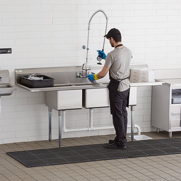 A man in a kitchen wearing gloves and an apron cleaning a Regency stainless steel 3 compartment sink.