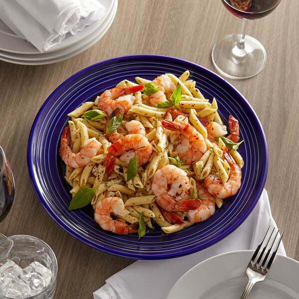 A Tuxton Concentrix cobalt blue china plate with pasta, shrimp, and basil on a table with a fork and a glass of wine.