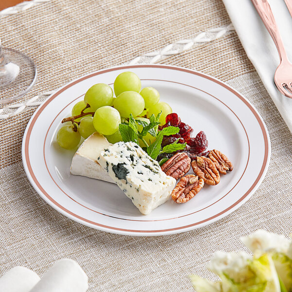 A Visions white plastic plate with rose gold bands holding cheese, grapes, and nuts on a table with other food plates.