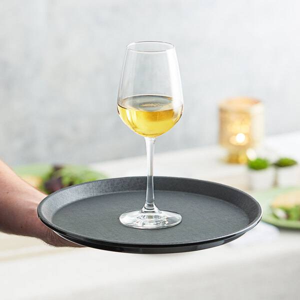 A person holding a Choice black non-skid serving tray with a glass of white wine.