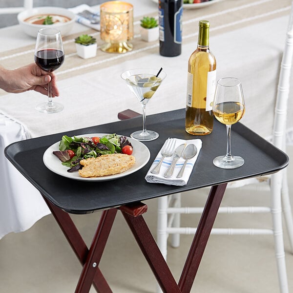 A person holding a Choice black non-skid serving tray with food and wine glasses on it.
