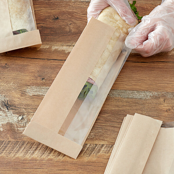 A person's hands in gloves packing a sandwich in a Choice brown Kraft bakery bag with a window.