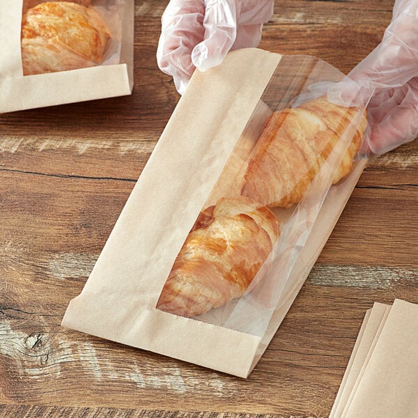 A person in gloves holding a Kraft window bag with a croissant inside.