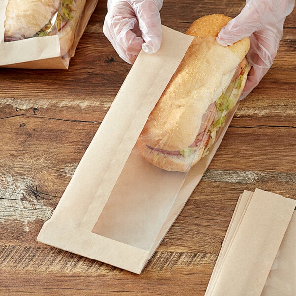 A person in gloves holding a sandwich in a Kraft paper bag with a window.