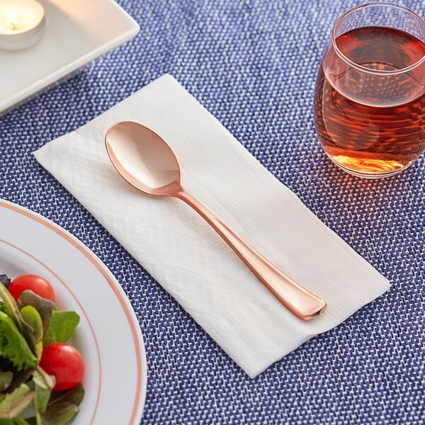 A Visions rose gold plastic spoon on a napkin next to a plate of salad and a glass of liquid.