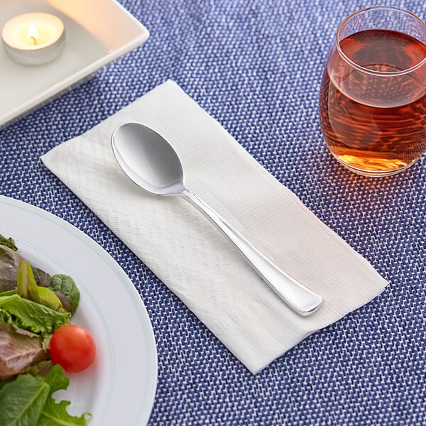 A Visions heavy weight silver plastic spoon on a napkin next to a plate of salad and a glass of liquid.