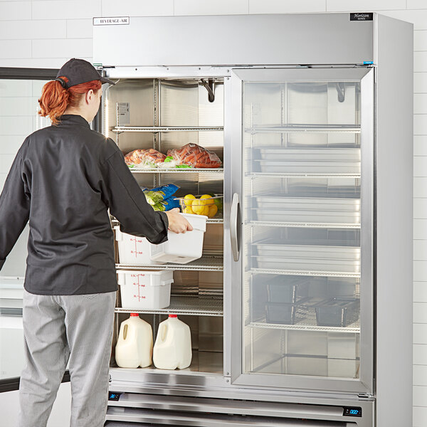A woman opening the glass door of a Beverage-Air stainless steel refrigerator in a professional kitchen.