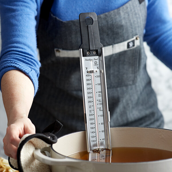 A person using a Comark candy/deep fry paddle thermometer in a pot of brown liquid.
