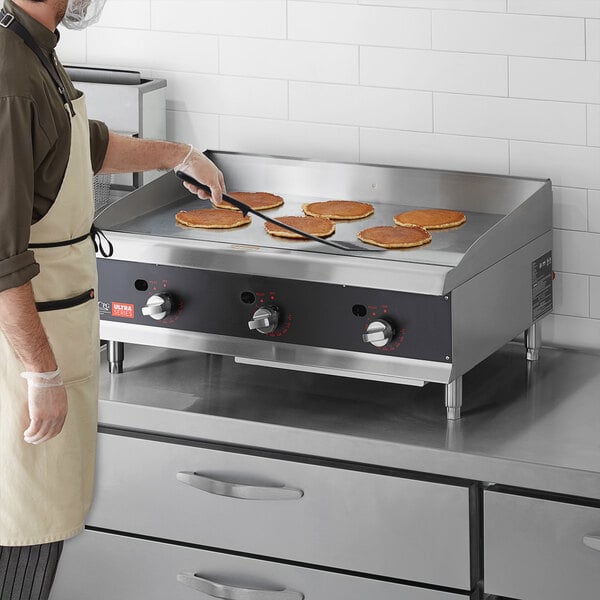 A man wearing a brown shirt and apron cooking pancakes on a Cooking Performance Group liquid propane countertop griddle.