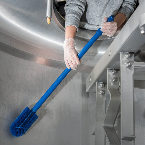 A person in gloves using a Carlisle blue brush to clean a stainless steel sink.