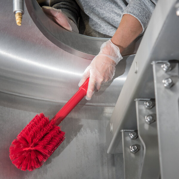 A person in white gloves using a red Carlisle Sparta multi-purpose brush to clean a metal surface.