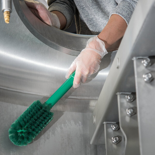 A person in gloves cleaning a circular stainless steel object with a green Carlisle Sparta brush.