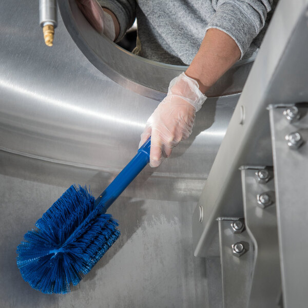 A person in gloves cleaning a metal cylinder with a blue Carlisle Sparta brush.