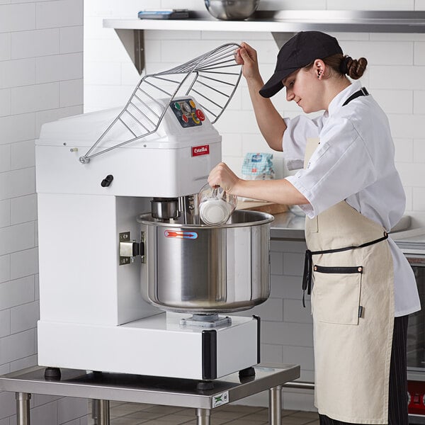 A woman in a chef's uniform using an Estella spiral dough mixer to mix a white liquid in a large metal bowl.
