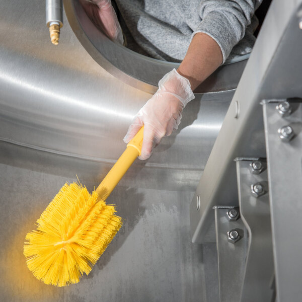 A person wearing gloves and holding a yellow Carlisle Sparta multi-purpose cleaning brush.