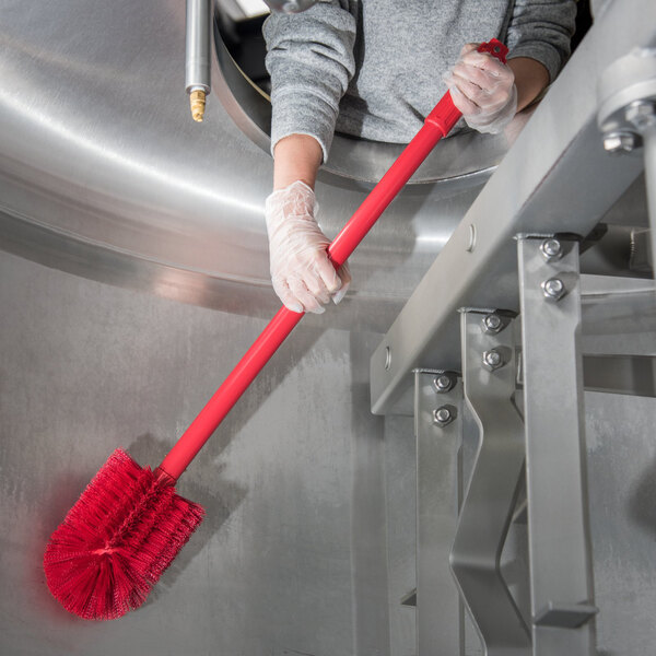 A person in gloves using a Carlisle red multi-purpose cleaning brush with a red handle to clean a metal container.