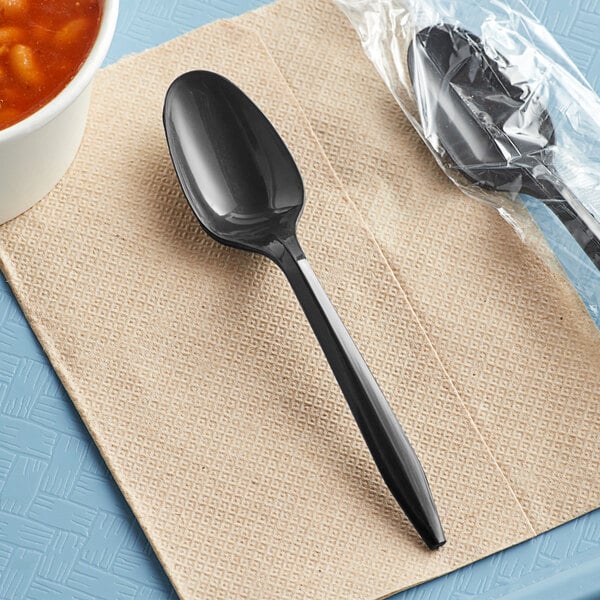 A black Choice plastic teaspoon on a napkin next to a bowl of soup.