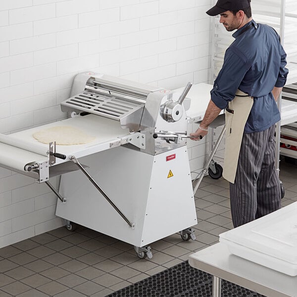 A man wearing a blue shirt and white apron standing by an Estella dough sheeter with dough on it.