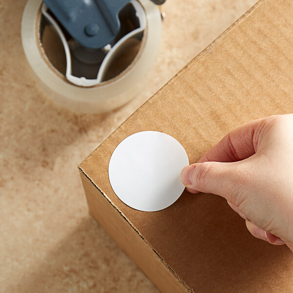 A hand using a Lavex round white inventory label on a cardboard box.