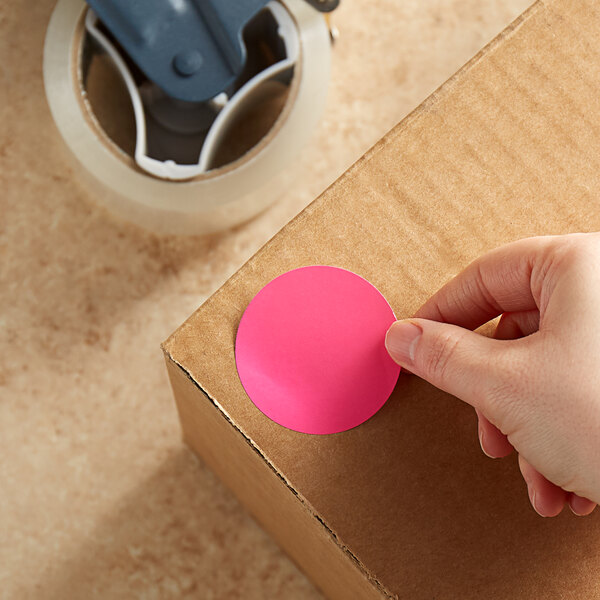 A hand using a Lavex fluorescent pink round sticker on a cardboard box.